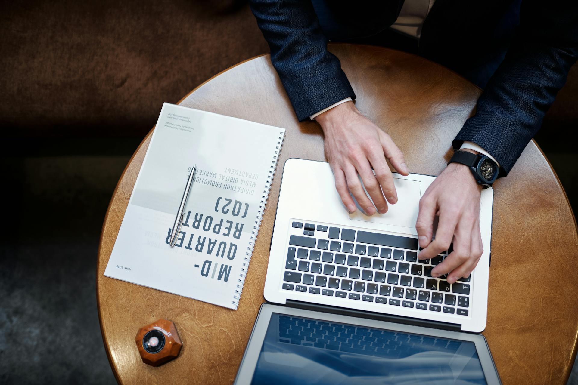 Top view of a businessman typing on a laptop next to a quarterly report on a wooden table.