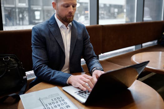 A professional businessman in a suit working on a laptop inside a cafe setting, focusing on a report.