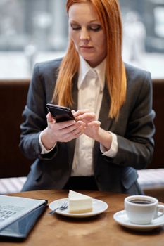 Professional woman in business attire using smartphone at a cafe with coffee and cake.