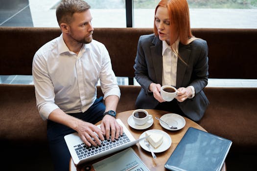 Two business professionals discussing work over coffee and using a laptop indoors.