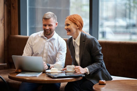 Two professionals in a café enjoying coffee and discussing work on a laptop.