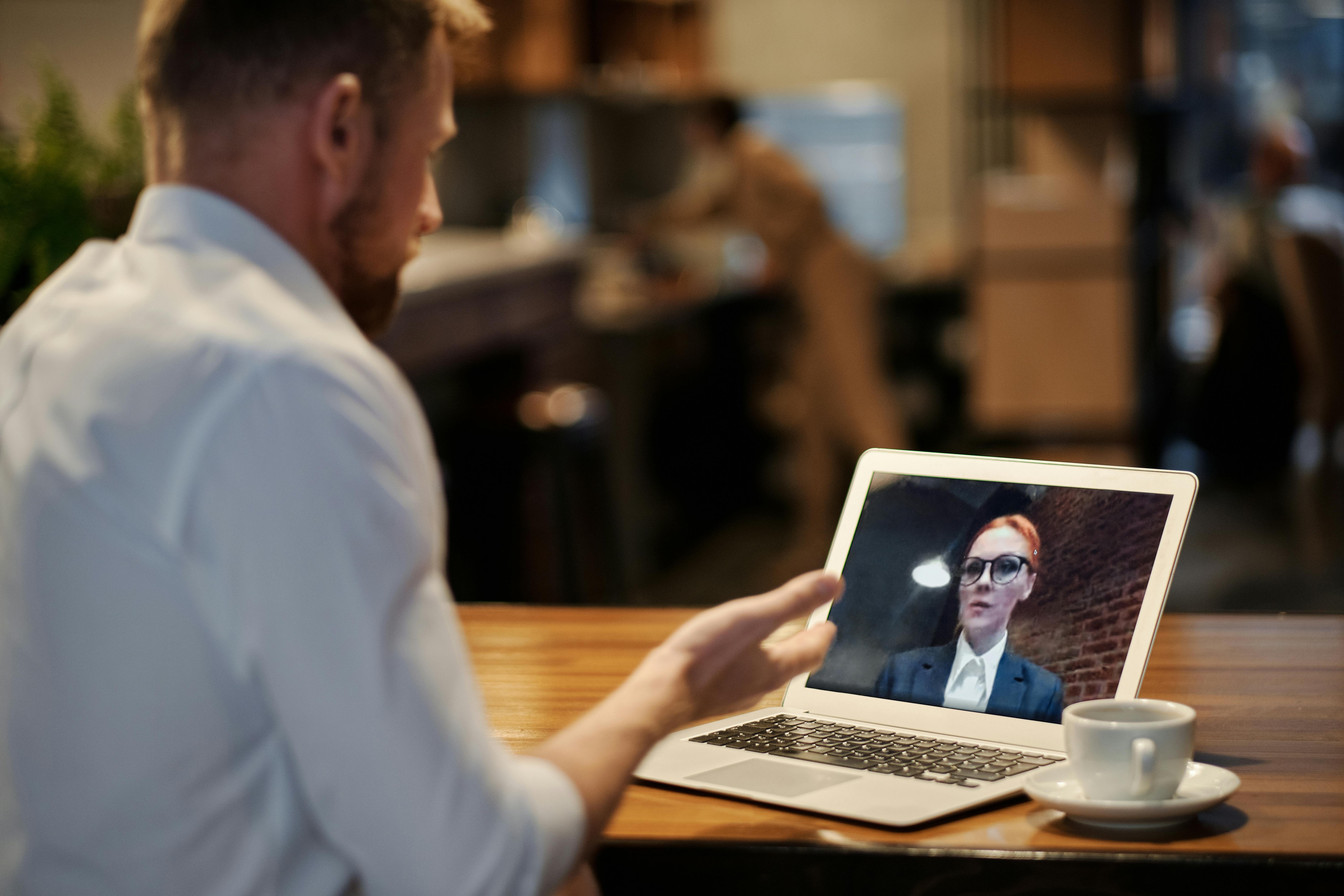 A man participates in an online business meeting via laptop in a warm, cozy coffee shop setting.