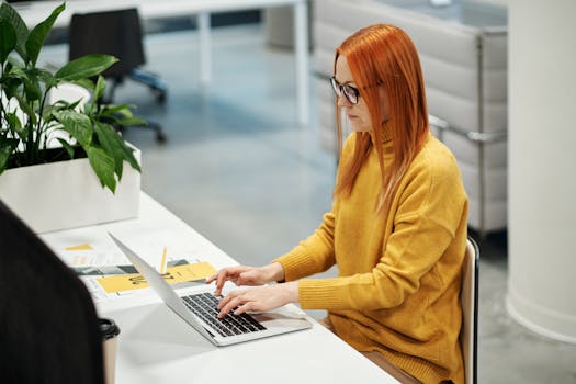 A woman in a mustard sweater working on her laptop in a modern office setting, surrounded by paperwork and plants.