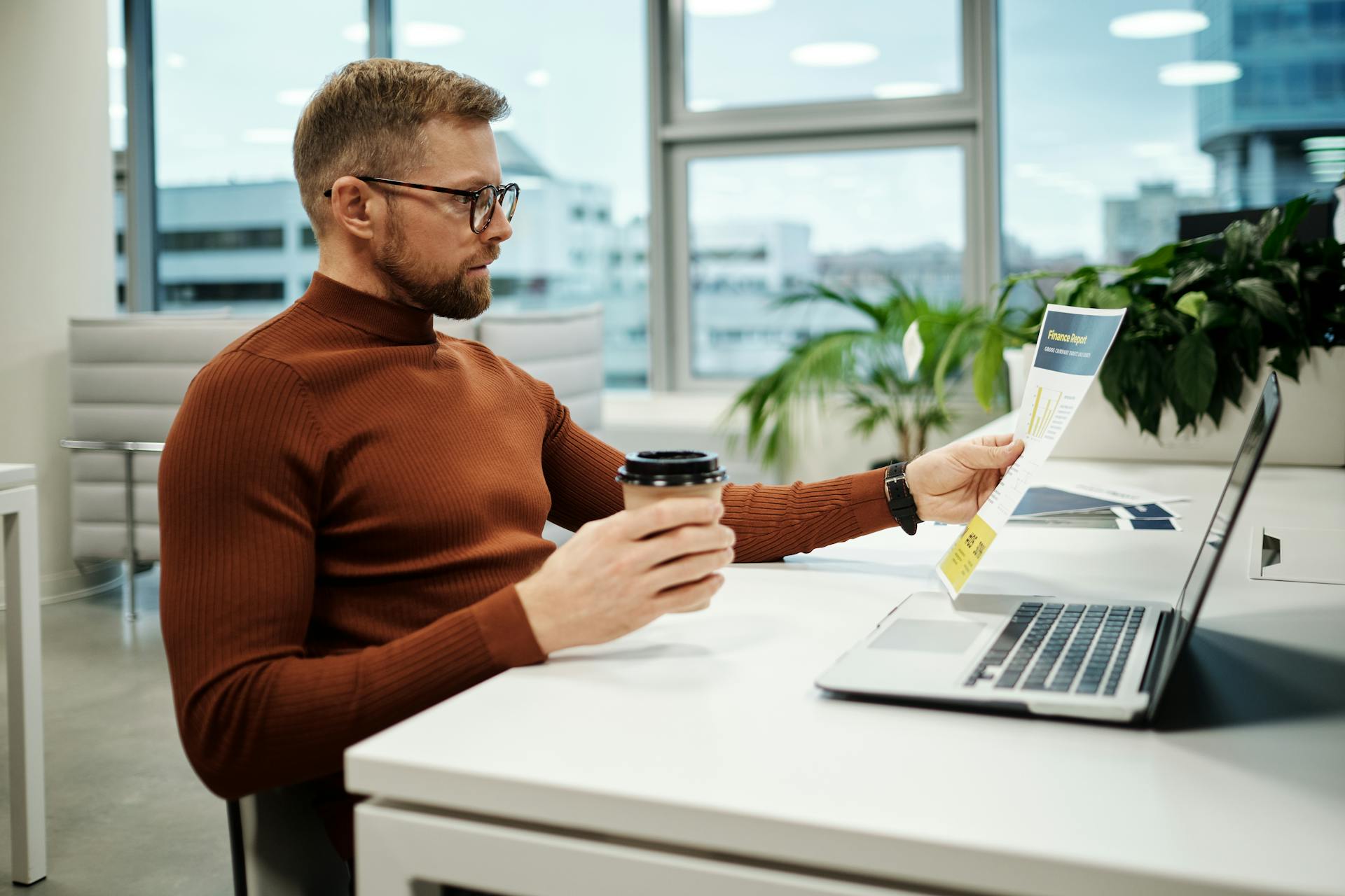 Professional businessman in brown turtleneck analyzing financial report while holding a takeaway coffee in modern office.