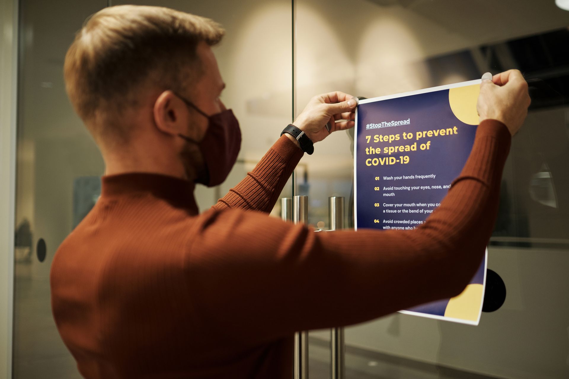 A man wearing a mask posts a COVID-19 prevention poster on a glass door indoors.