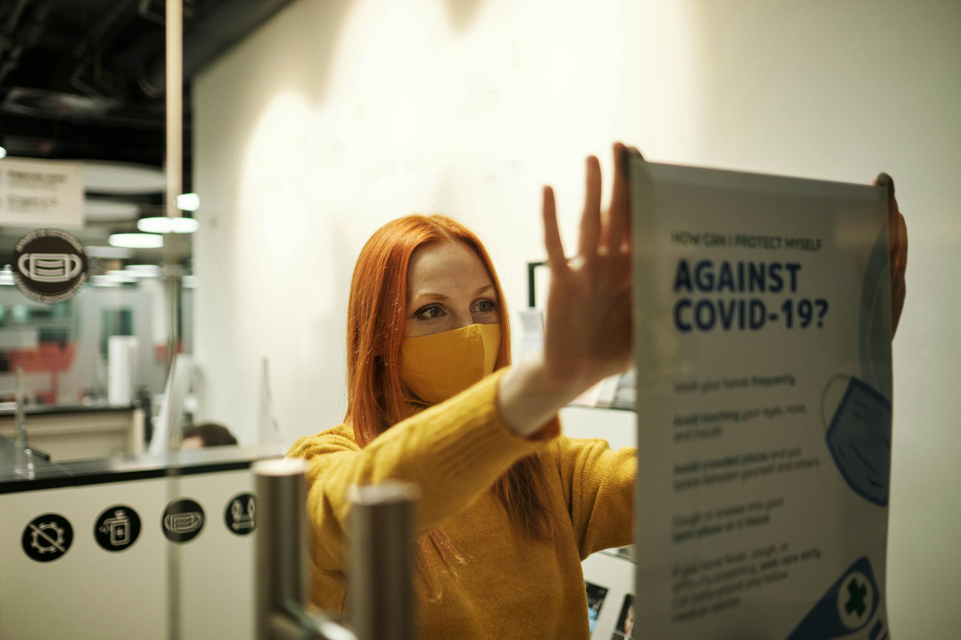 A woman in a yellow sweater hangs a COVID-19 safety poster in an office setting.