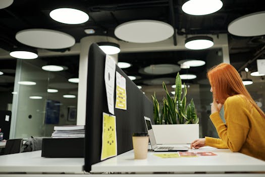 Red-haired woman concentrating on laptop work in a stylish office with plants and coffee.