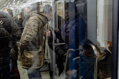 Through glass view of men surfing internet on cellphones against crop anonymous people during trip in underground public transport