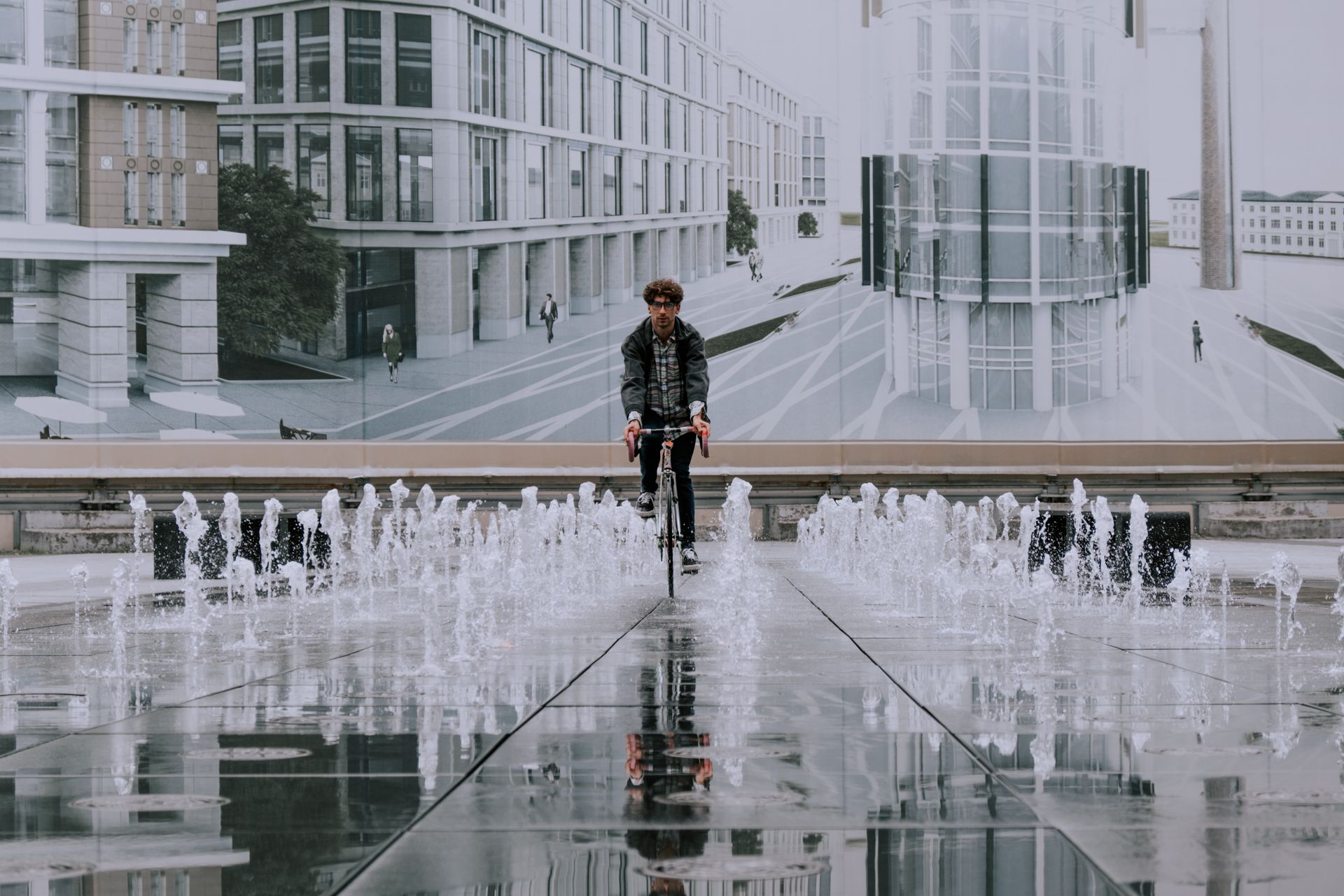 A man cycles through an urban area with modern architecture and reflecting fountains on wet pavement.