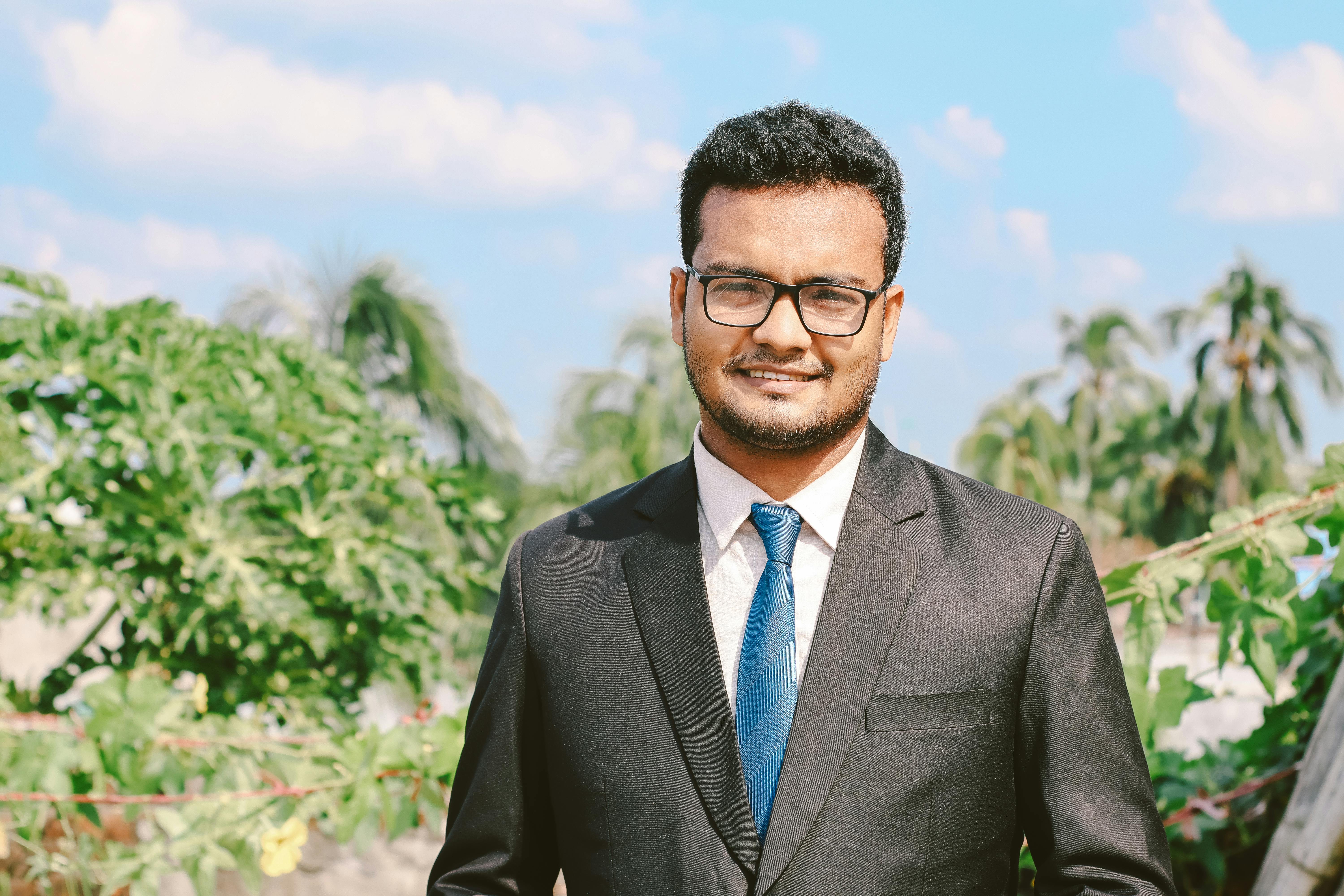 Portrait of a smiling man in a suit and eyeglasses, exuding confidence outdoors with a clear blue sky.