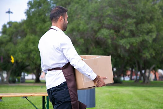 Man in uniform carries a box outdoors in a lush park setting, depicting a delivery scene.