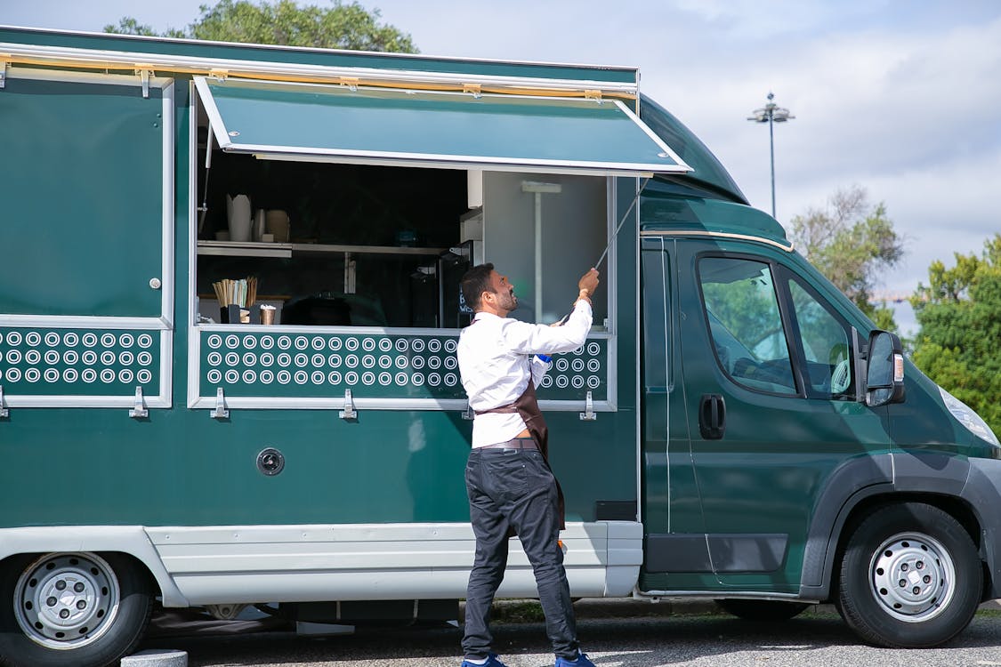 Side view young waiter in apron standing outside food truck and opening counter door in green lush park