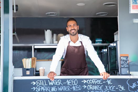 Good looking young waiter in apron standing at modern food truck counter and looking at camera with toothy smile