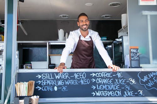 Low angle of ethnic seller wearing uniform standing at counter in food trailer and smiling happily