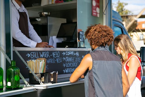 Multiethnic couple standing together near food truck with seller and selecting dishes from menu in sunny day