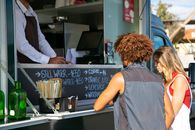 Multiethnic couple standing together near food truck with seller and selecting dishes from menu in sunny day