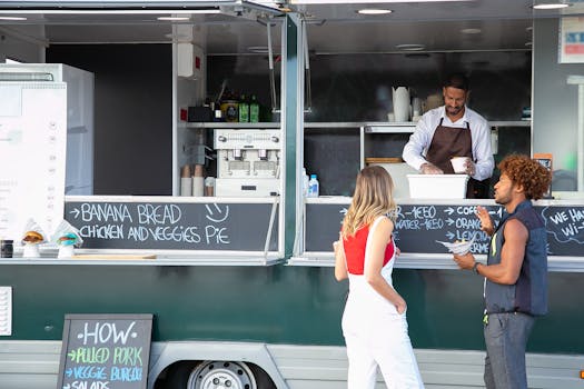 Two people order food from a vendor at a street-side food truck in daylight.