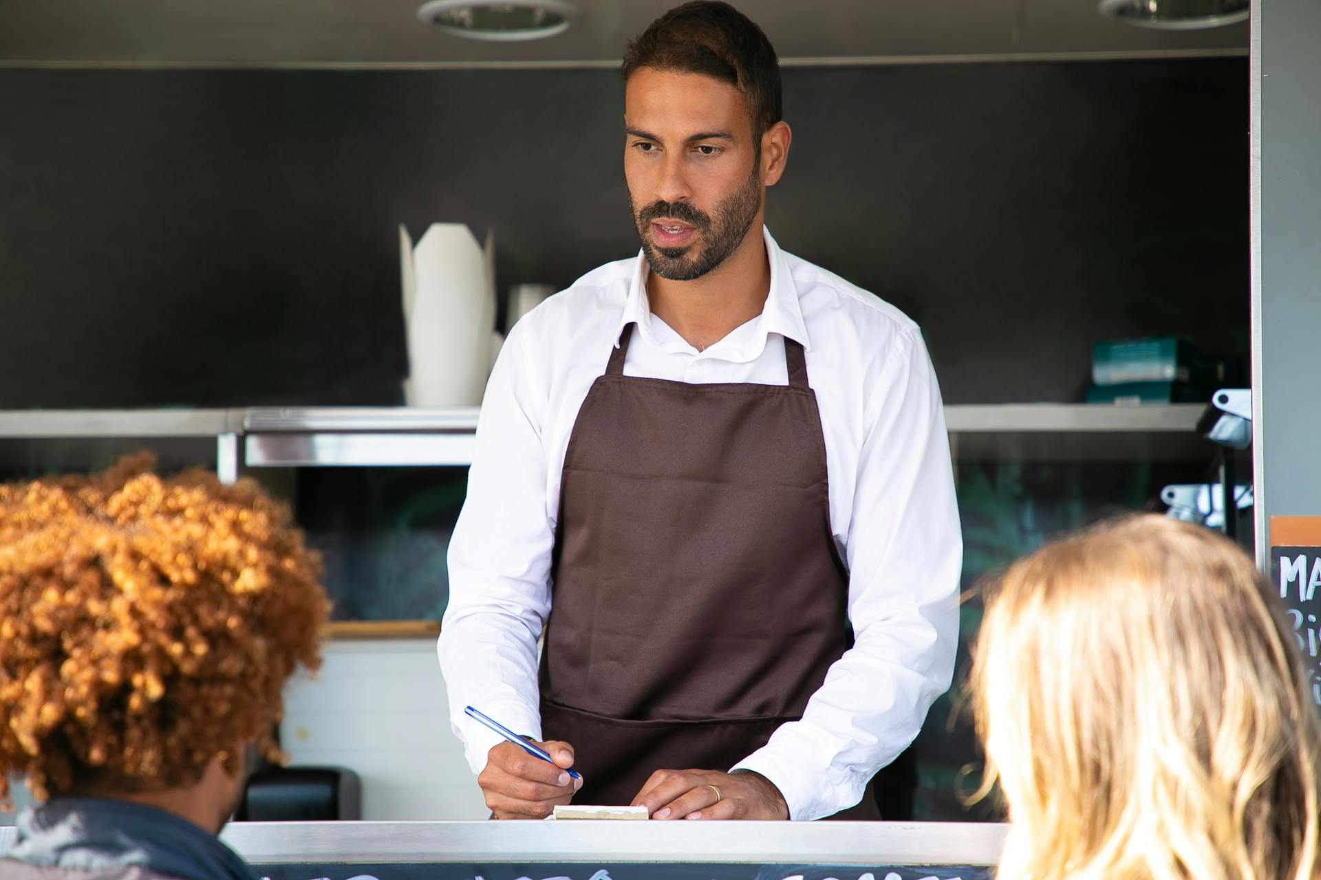 Ethnic male seller talking to client and writing order on paper while working in street food truck