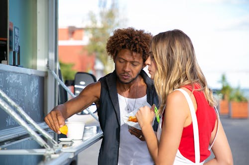 Free Hungry young black man and woman in casual clothes eating yummy crisps with sauce and hamburger while having lunch in food truck Stock Photo
