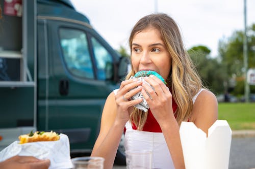 Young hungry female in casual clothes eating tasty hamburger and looking away in street cafe near food truck on sunny day