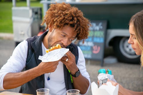 Hungry young African American male with curly hair in casual clothes eating tasty sandwich while spending time with crop girlfriend in street cafe in outskirts