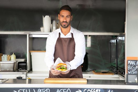Concentrated young ethnic male cook in white shirt and apron serving appetizing hamburger during work in food truck
