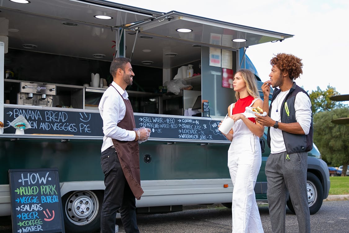 Side view of happy young ethnic waiter in uniform standing near food van and smiling while greeting diverse customers eating delicious takeaway burger and french fries
