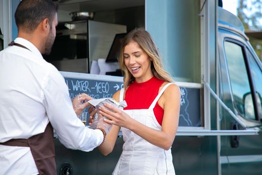 Crop anonymous waiter giving tasty burger to happy young female client standing near van cafe in daytime