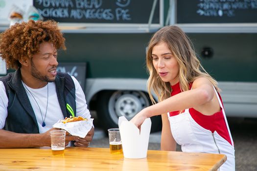 Positive young female eating french fries and drink beer while sitting at table near black boyfriend with burger in hand in road cafe
