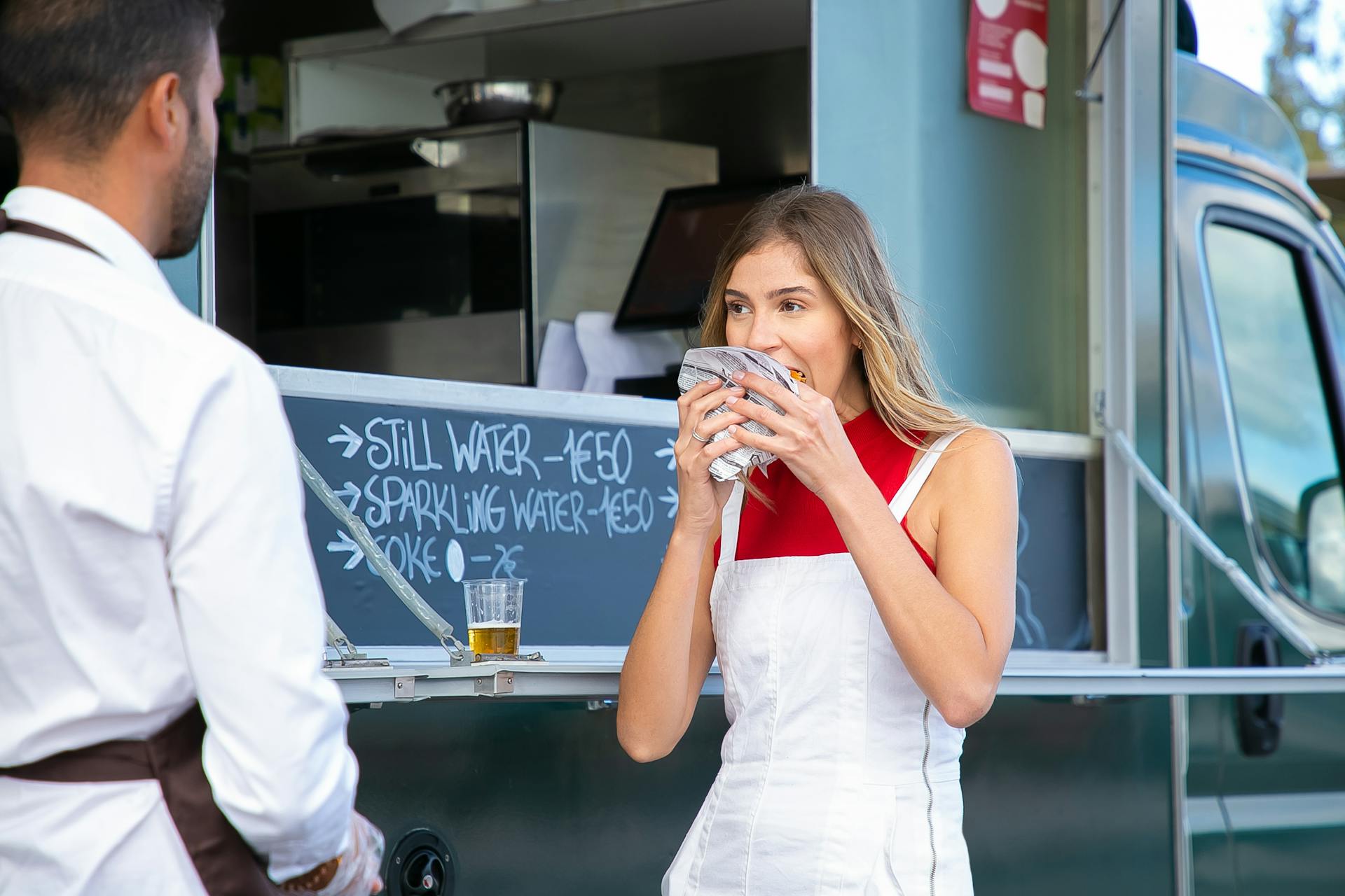 Calm woman in stylish clothes standing near food truck and eating tasty burger on street in daytime