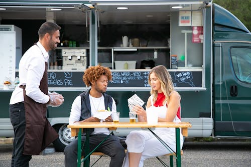 Cheerful owner of food truck standing near table with diverse couple eating burgers and resting on street in daytime