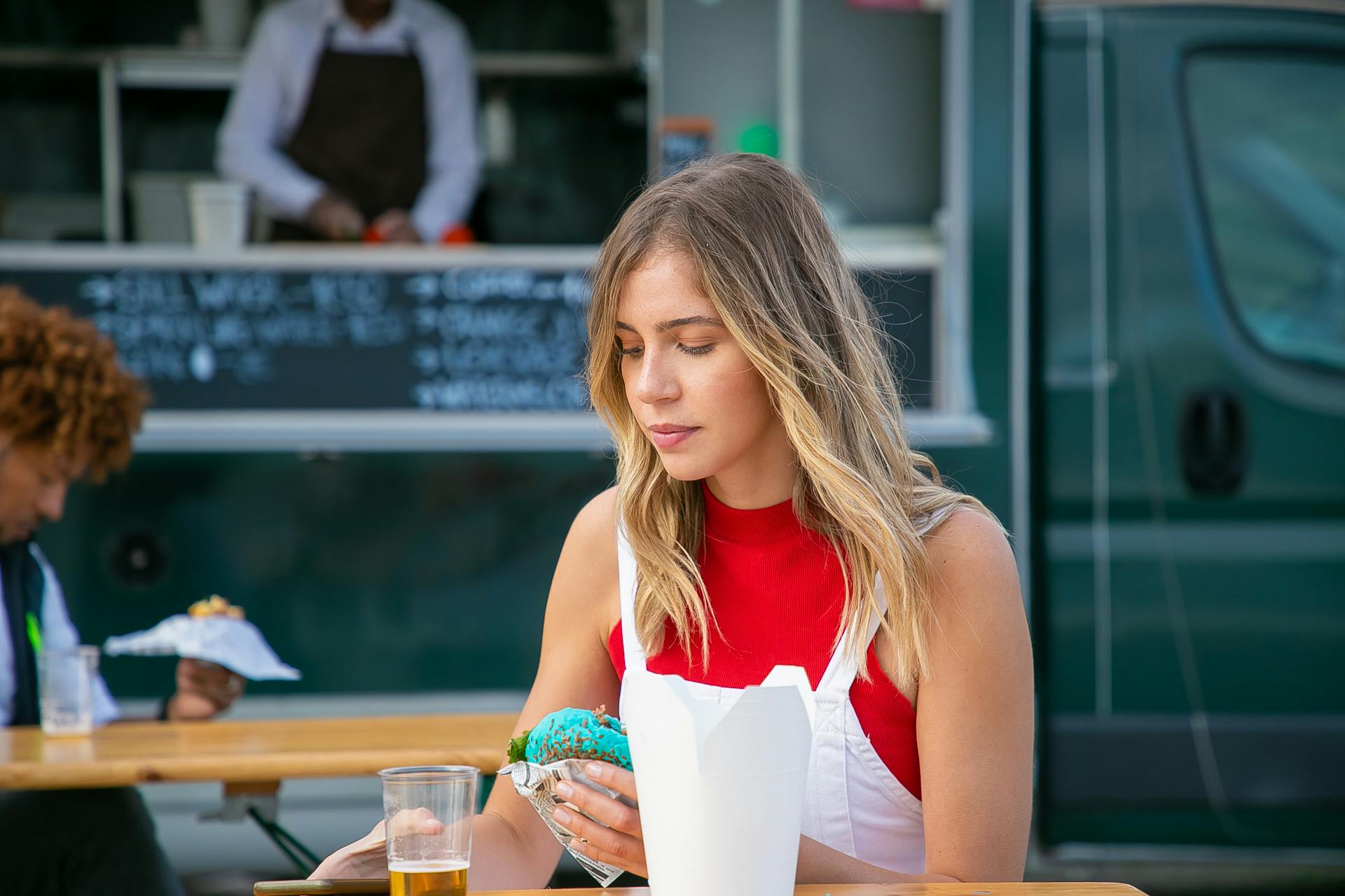 Tranquil woman with blond hair in casual clothes sitting at table with drink and eating blue burger in street cafe against food truck in daytime