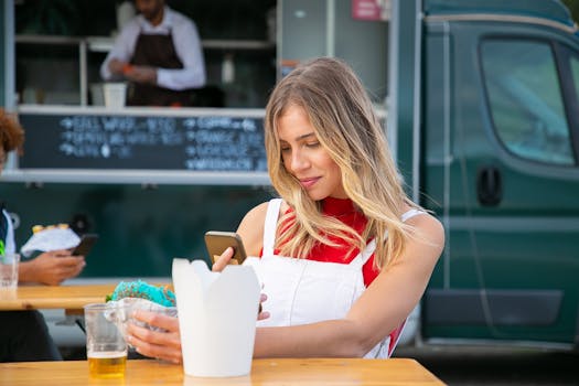 A cheerful young woman using a smartphone while having lunch at a food truck outdoors.
