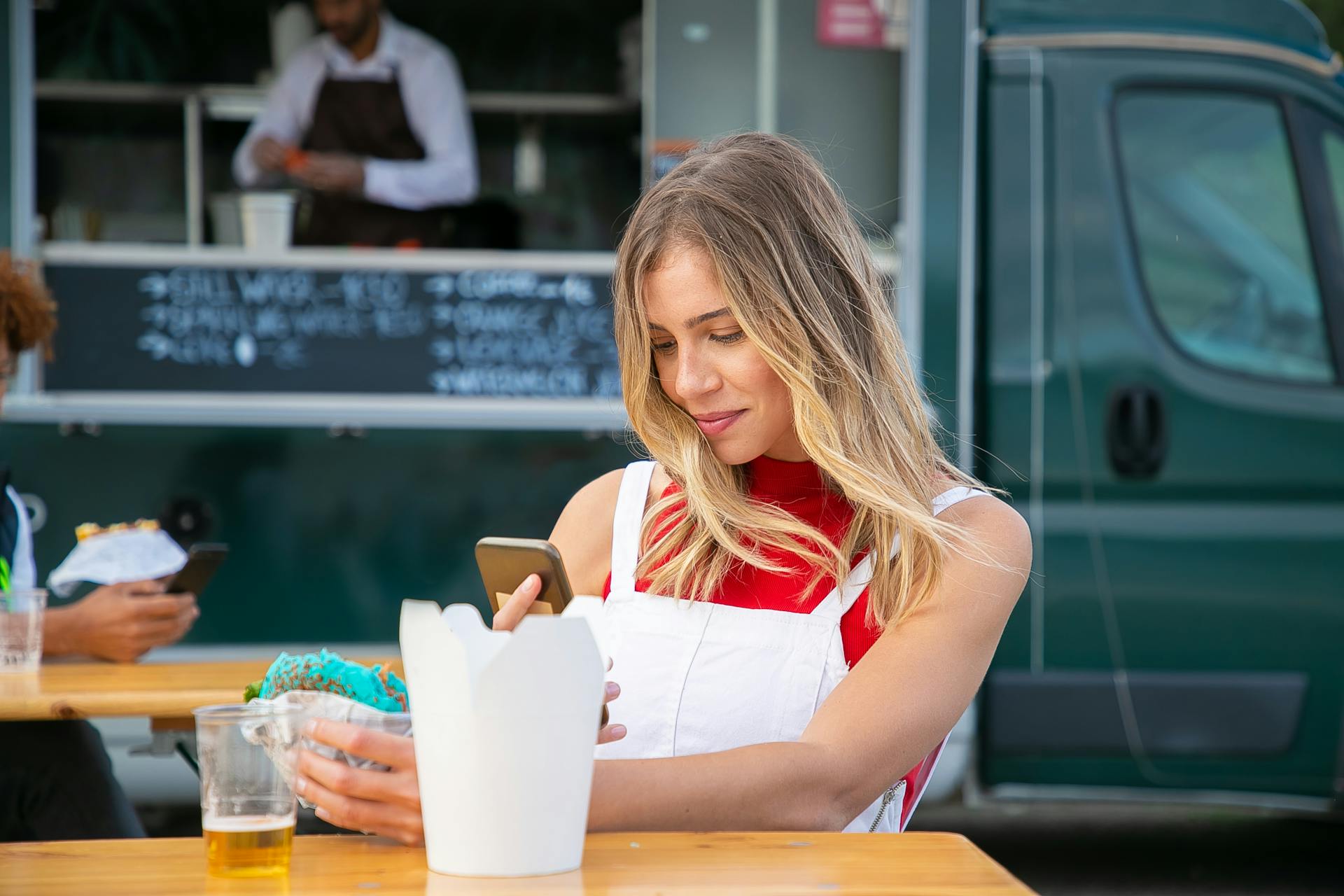 A cheerful young woman using a smartphone while having lunch at a food truck outdoors.