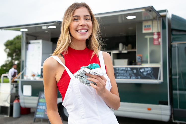 Happy Woman Standing With Burger Against Food Truck