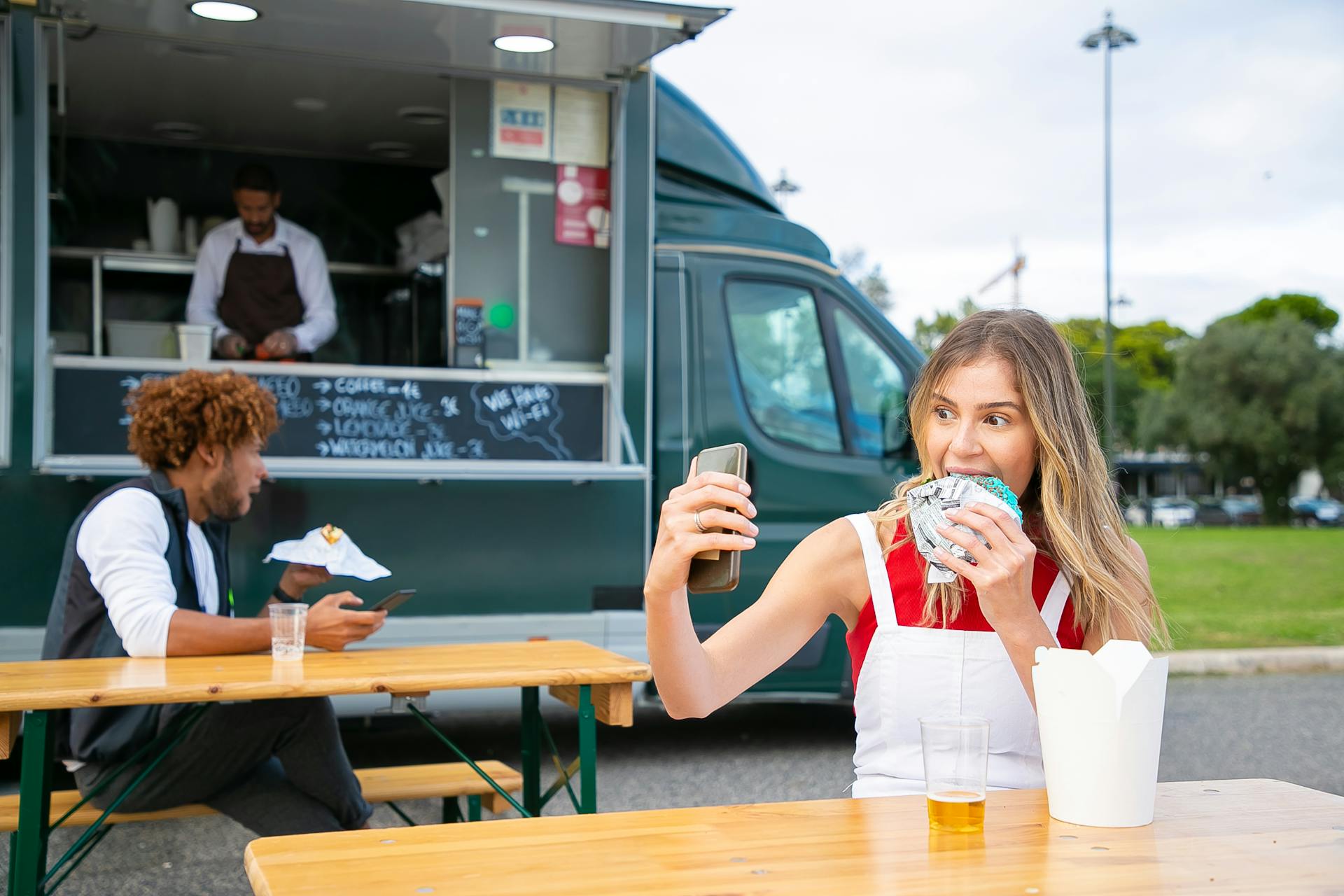 Cheerful young woman in casual clothes eating delicious blue burger and taking selfie while chilling on street near food truck with busy owner and black man eating hamburger in daytime
