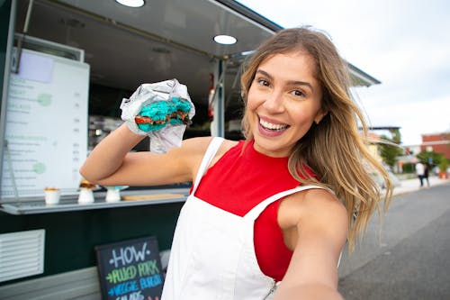 Smiling female in casual clothes standing with blue burger and taking selfie on street against food truck in daytime