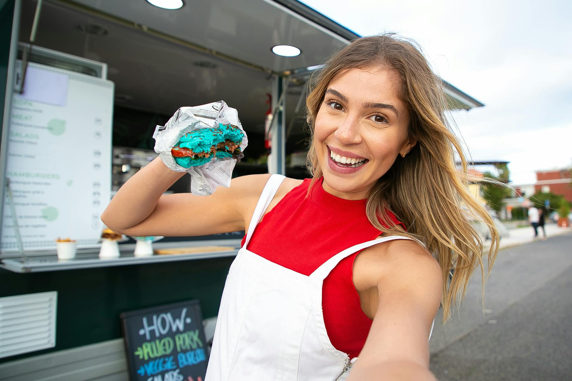 Smiling female in casual clothes standing with blue burger and taking selfie on street against food truck in daytime