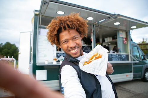 Cheerful African American man with curly hair in casual clothes taking selfie with fresh burger against food truck in daytime
