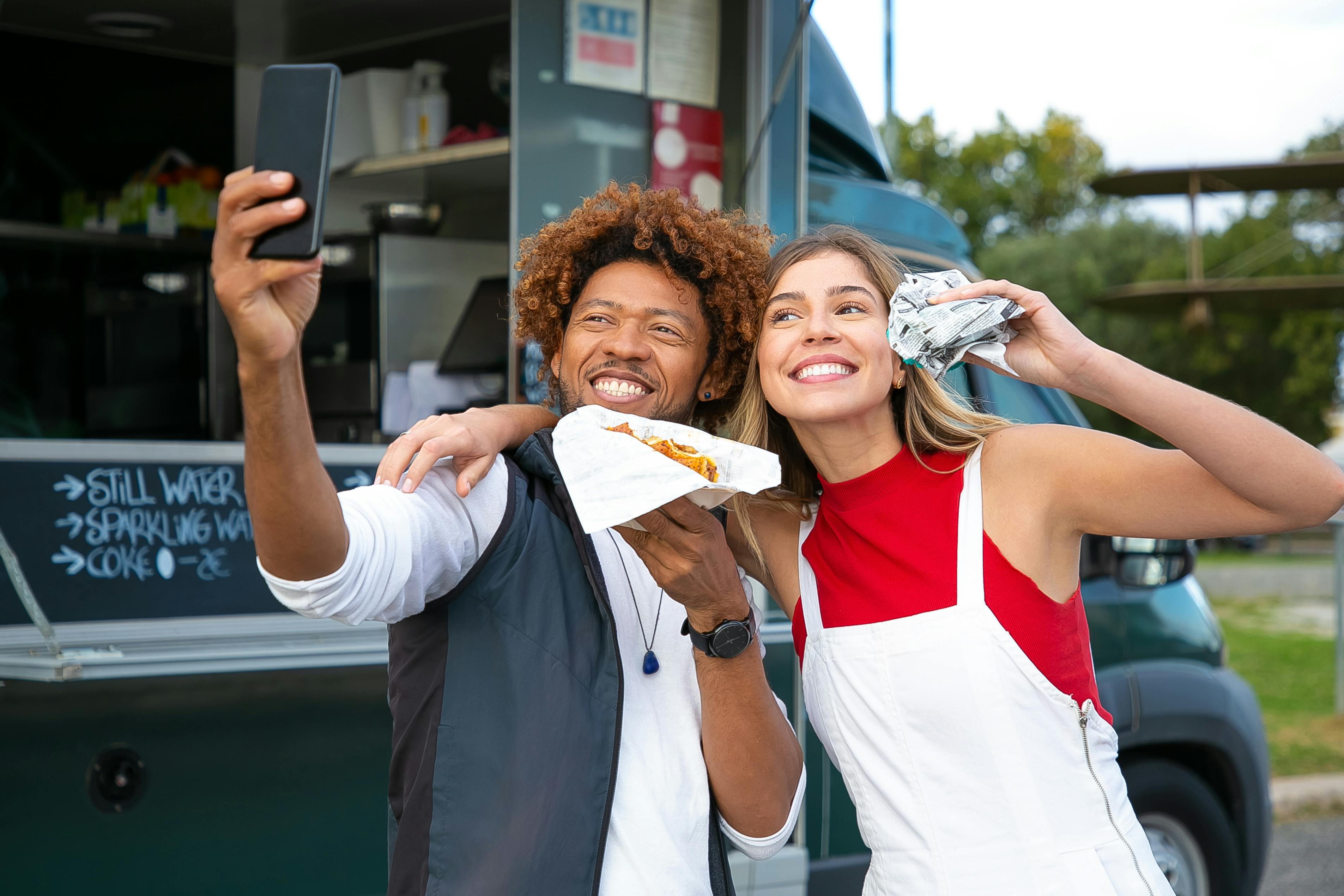 happy multiethnic friends with burgers taking selfie near food truck