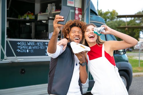 Joyful multiracial friends embracing and taking selfie on cellphone while eating tasty burgers near food truck in lush park