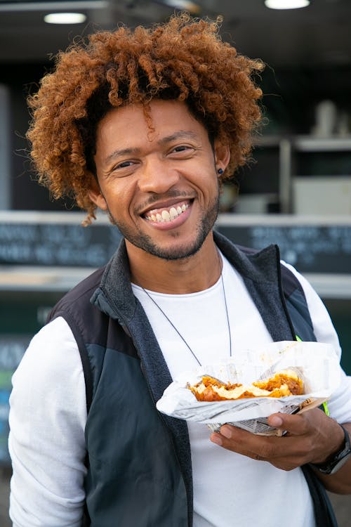 Cheerful young African American male with curly hair enjoying yummy sandwich near street food truck and looking at camera with toothy smile