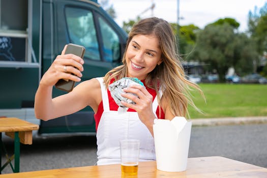 A cheerful young woman enjoys street food outdoors while taking a selfie, capturing a joyful moment.