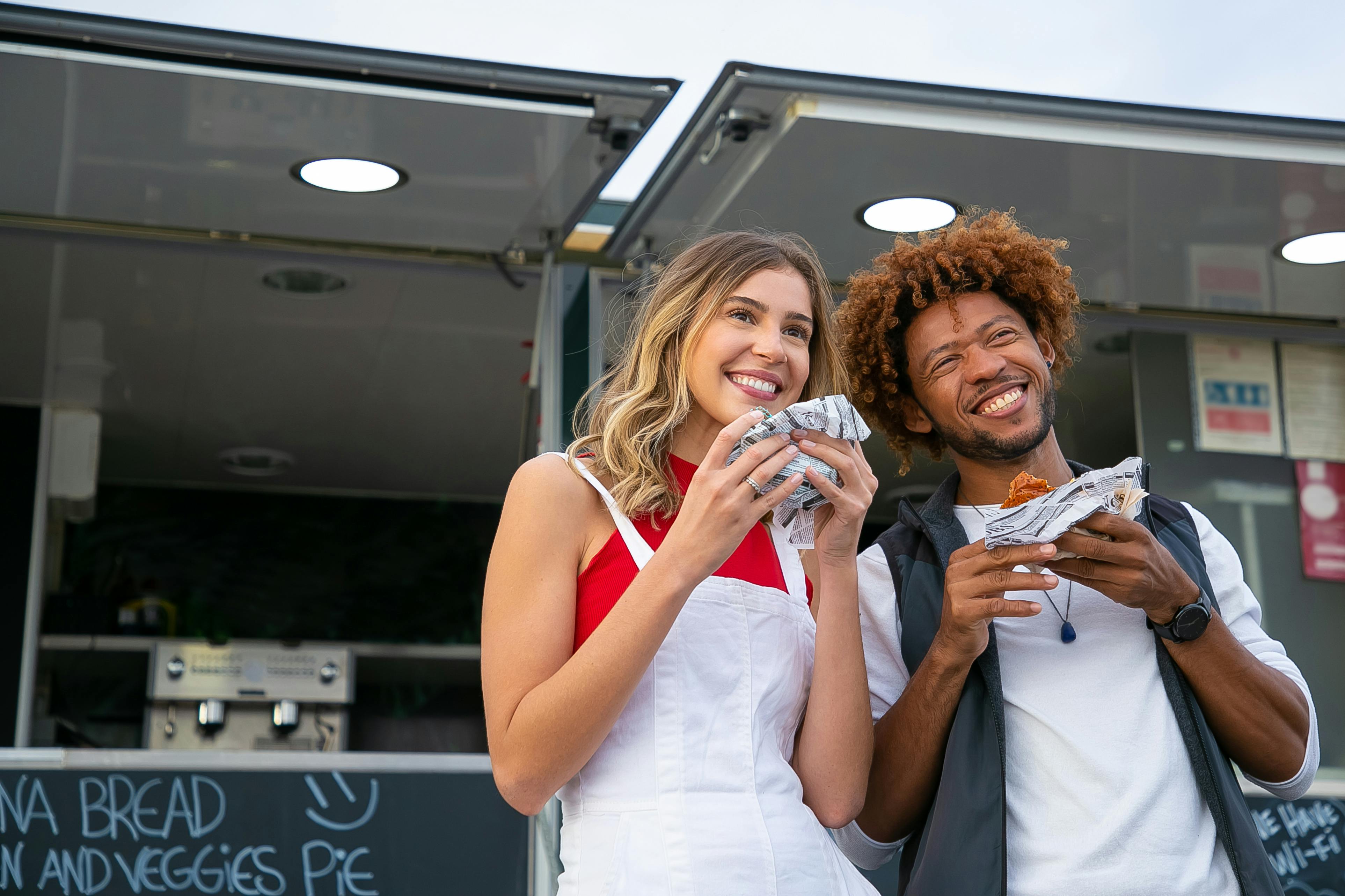 Delighted young multiracial friends wearing casual outfits enjoying delicious burgers and looking away with smiles while standing near street food truck