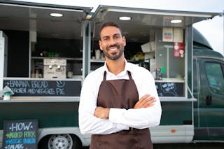 Smiling young ethnic male waiter in apron standing with arms folded near modern food truck and looking at camera contentedly