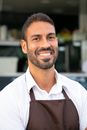 Cheerful ethnic waiter in apron standing against street cafe