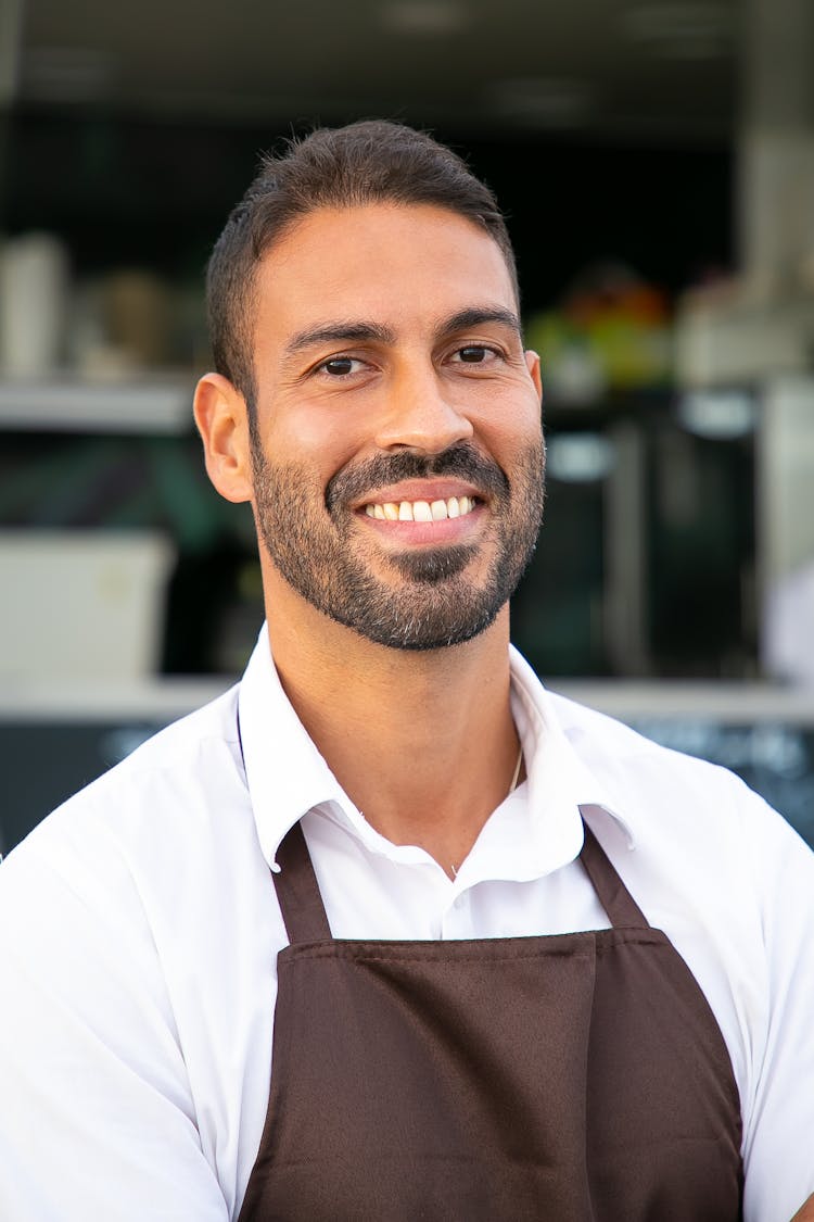 Cheerful Ethnic Waiter In Apron Standing Against Street Cafe