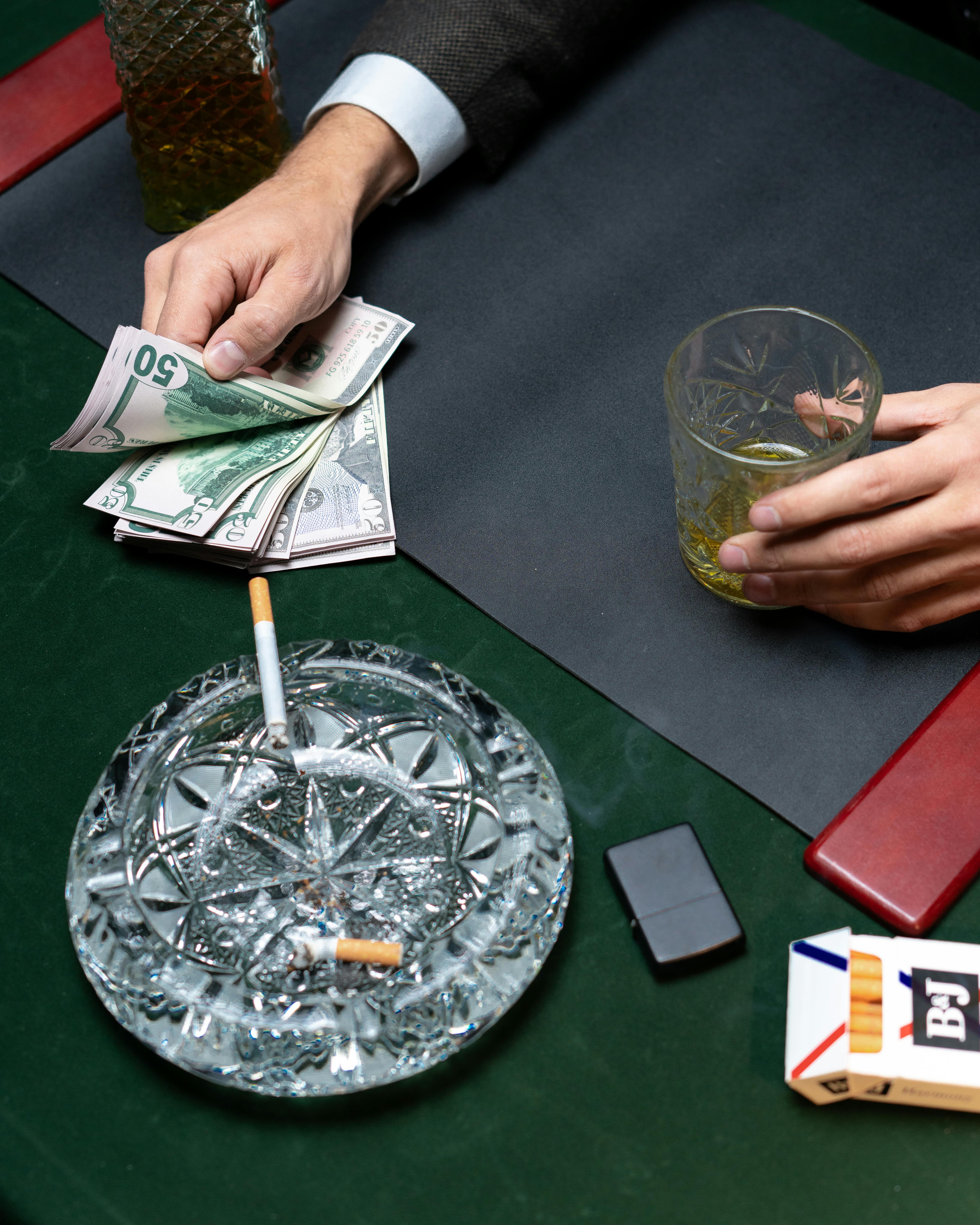 Close-up of a sophisticated table setting with whiskey, cash, and a lit cigarette.