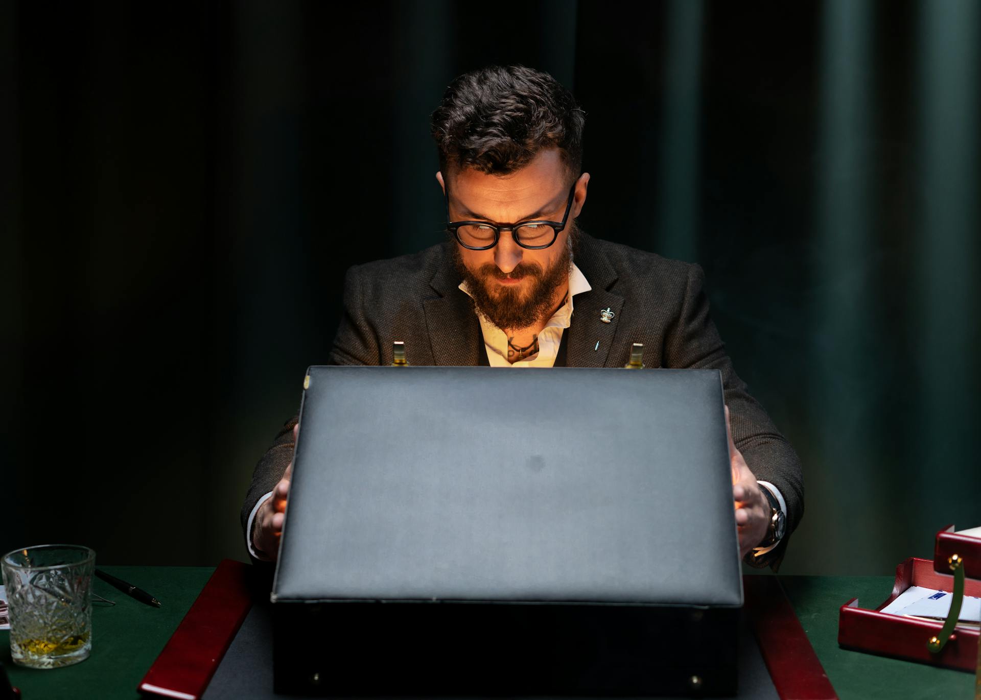 A bearded businessman wearing glasses opens a briefcase in a dimly lit office.