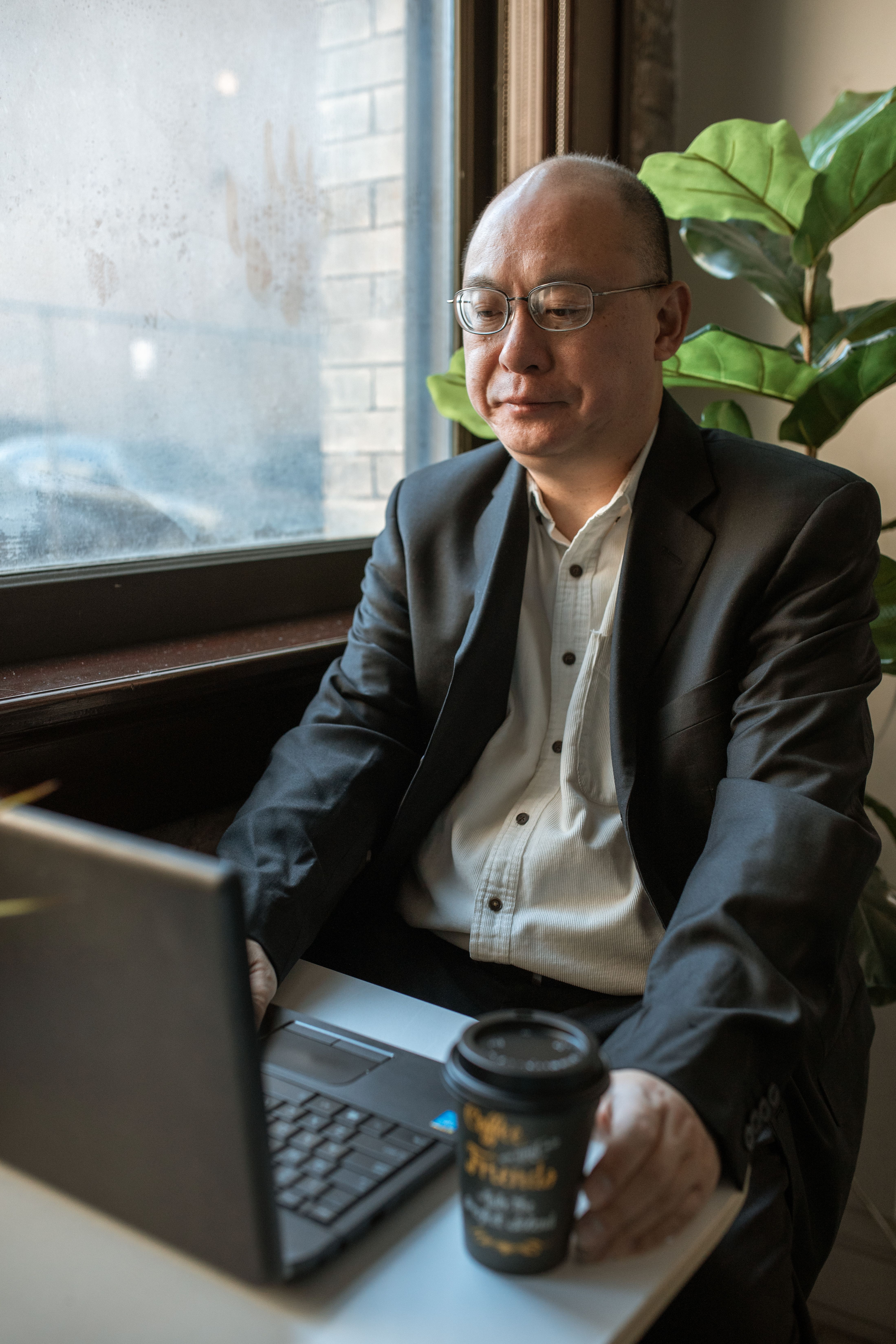 亚洲商人在一家舒适的咖啡馆里，对着窗户用笔记本电脑工作